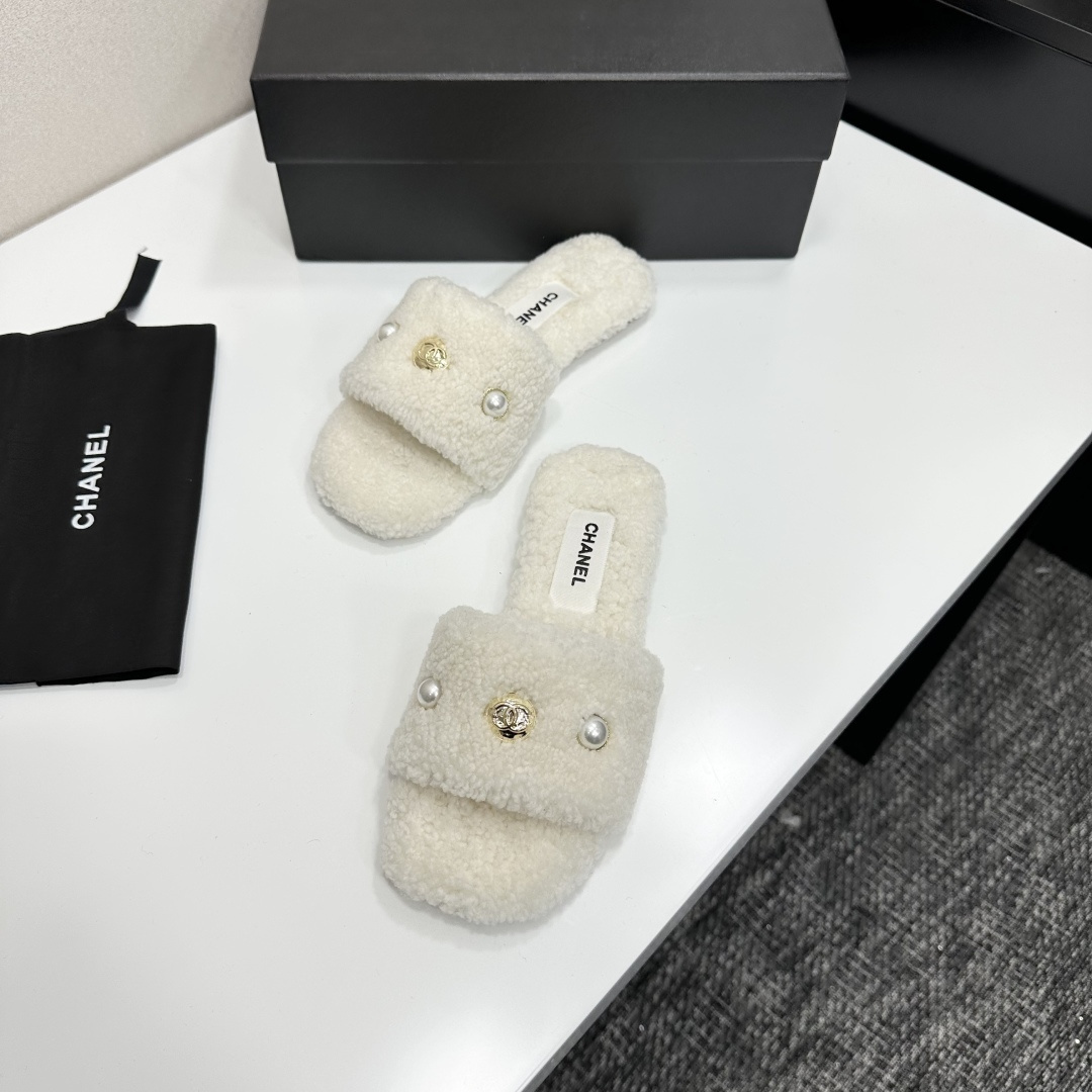 A pair of white Chanel shearling slippers featuring pearl and gold CC logo embellishments sitting on a black designer box. - Hình 5