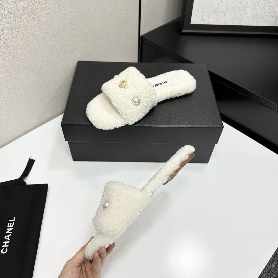 A pair of white Chanel shearling slippers featuring pearl and gold CC logo embellishments sitting on a black designer box. - Hình 3