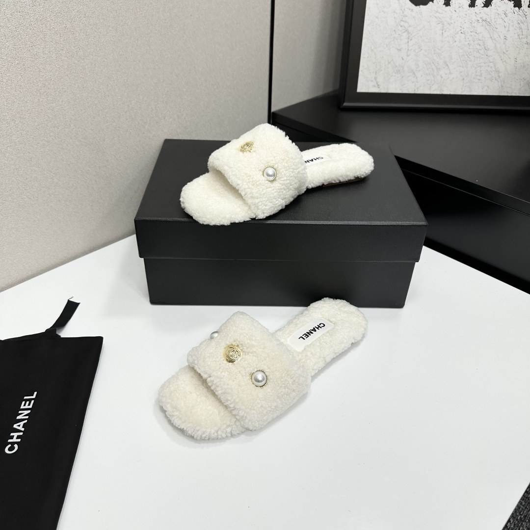 A pair of white Chanel shearling slippers featuring pearl and gold CC logo embellishments sitting on a black designer box. - Hình 2