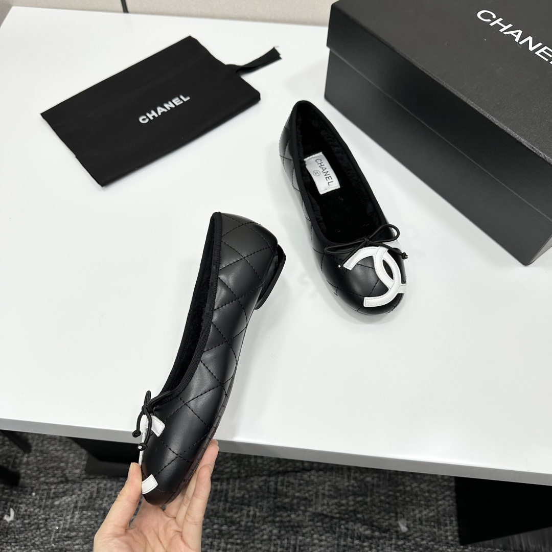 A pair of black quilted Chanel ballet flats with white CC logo on the toe and shearling lining, resting on a black box. - Hình 8