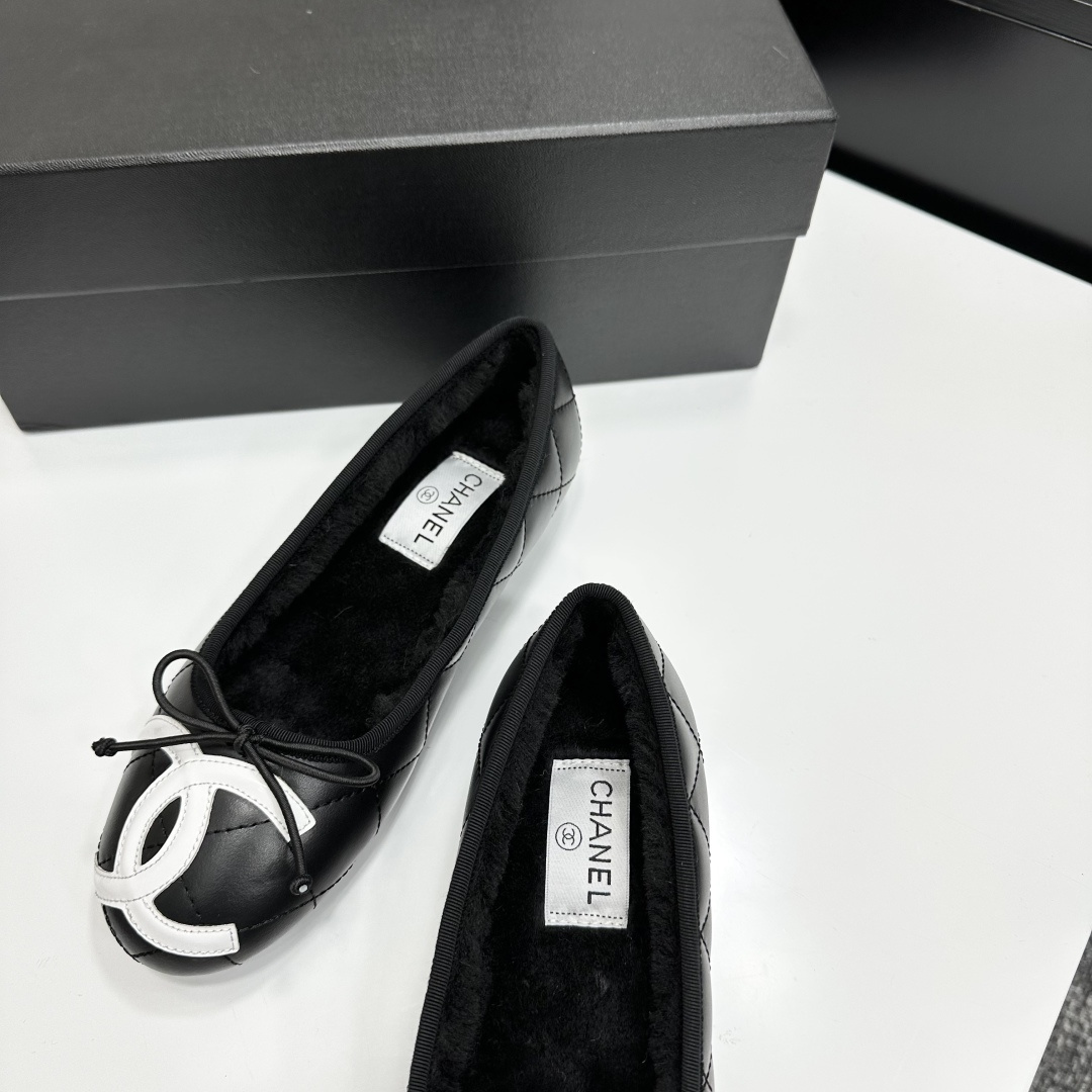A pair of black quilted Chanel ballet flats with white CC logo on the toe and shearling lining, resting on a black box. - Hình 6