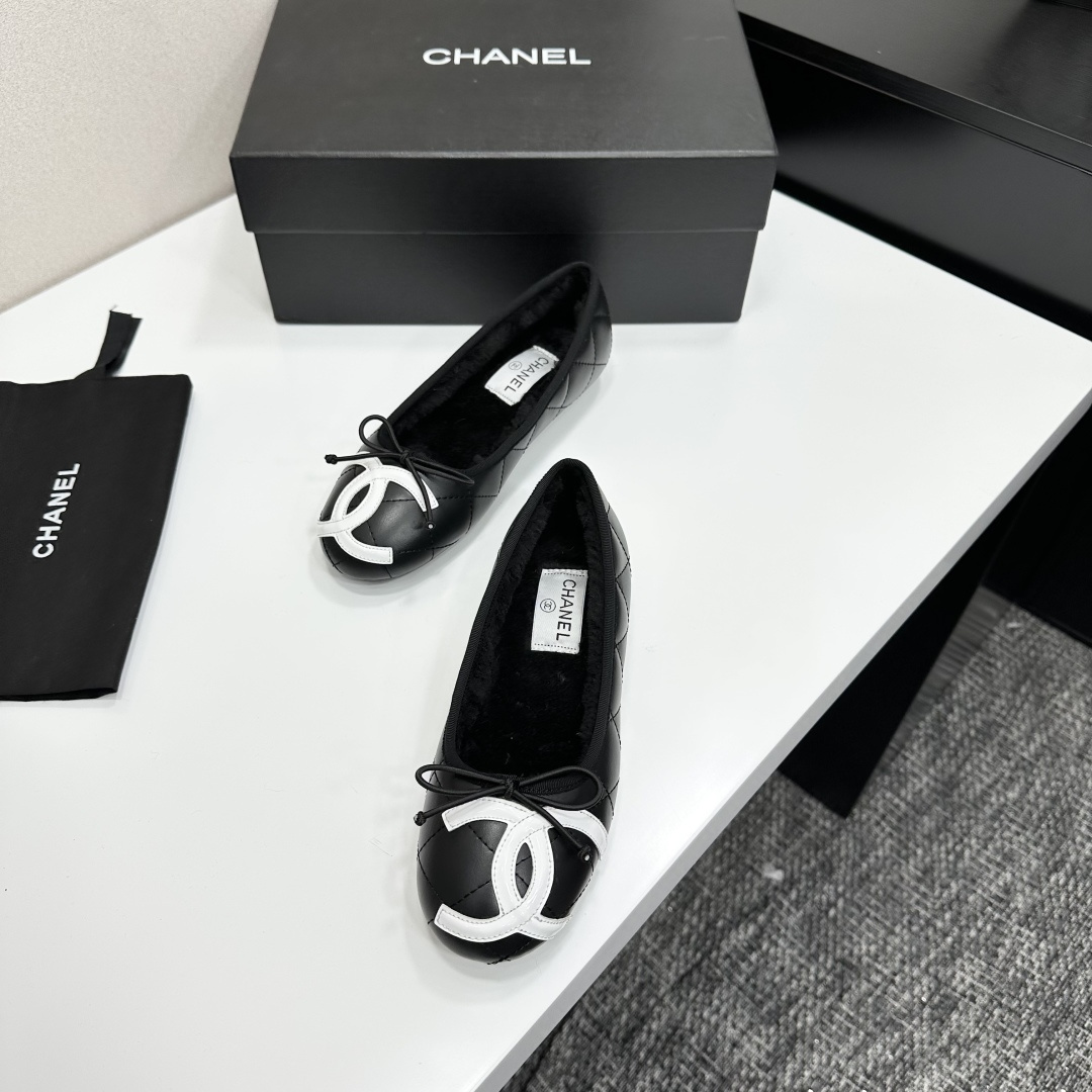 A pair of black quilted Chanel ballet flats with white CC logo on the toe and shearling lining, resting on a black box. - Hình 5