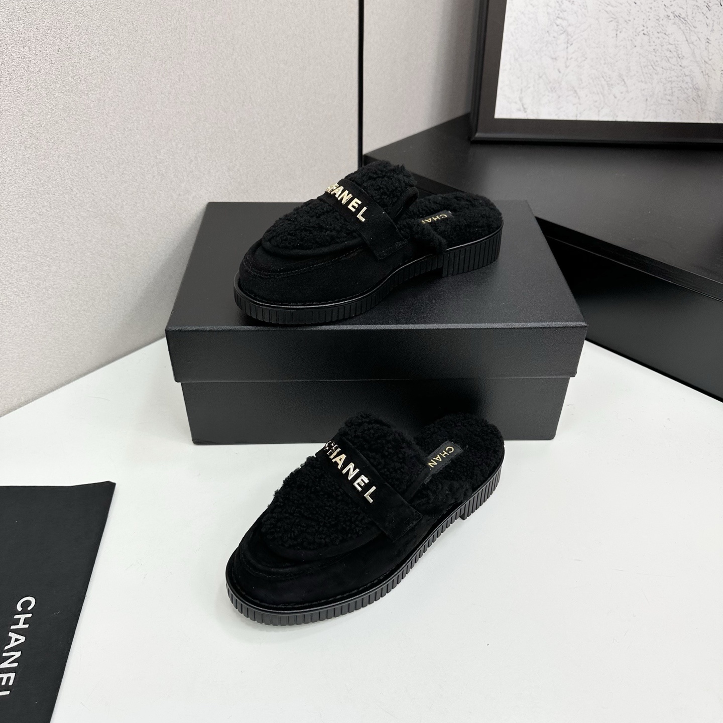 A pair of luxury Chanel black suede mules with fuzzy shearling lining and gold-tone logo hardware on a black platform sole. - Hình 2