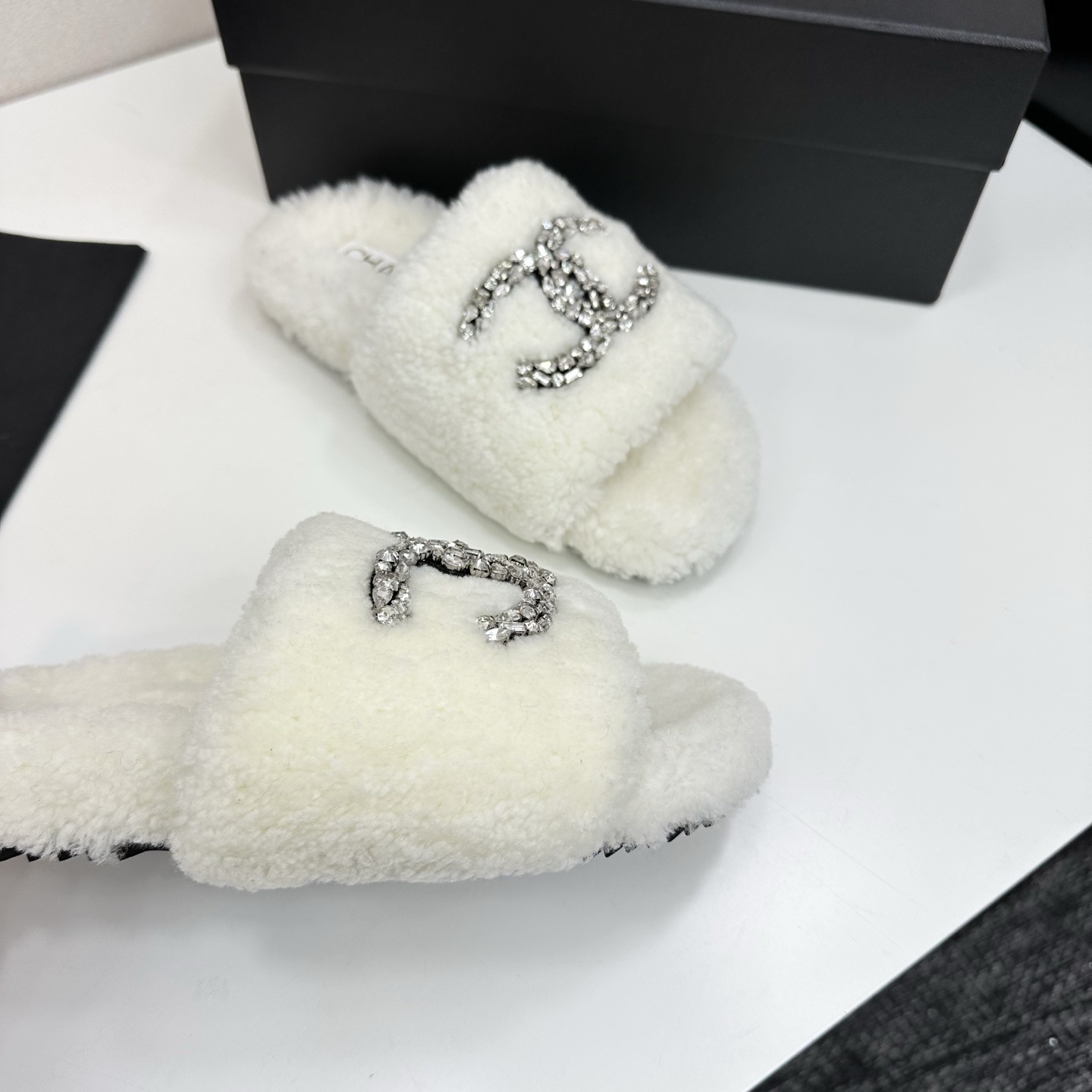 A pair of white fuzzy shearling slides with a crystal-embellished logo on a black box. - Hình 9