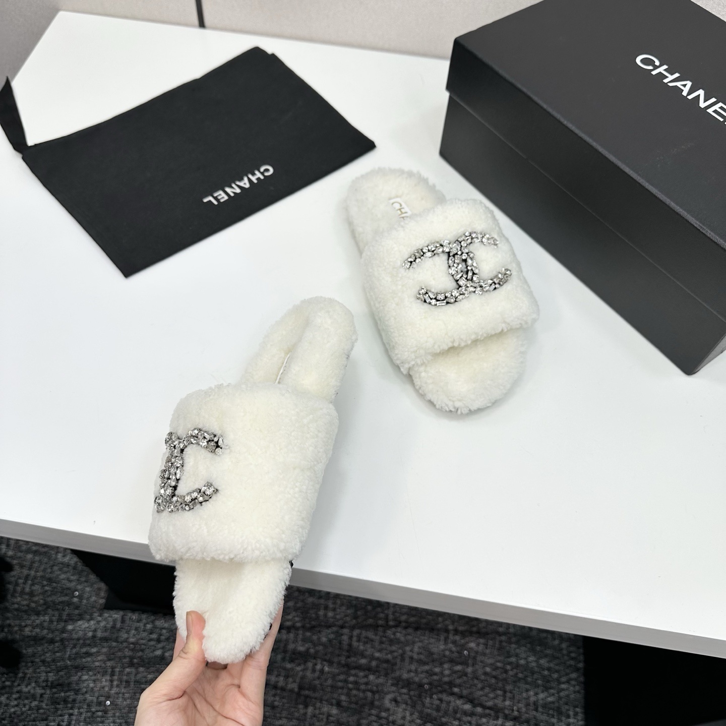 A pair of white fuzzy shearling slides with a crystal-embellished logo on a black box. - Hình 8