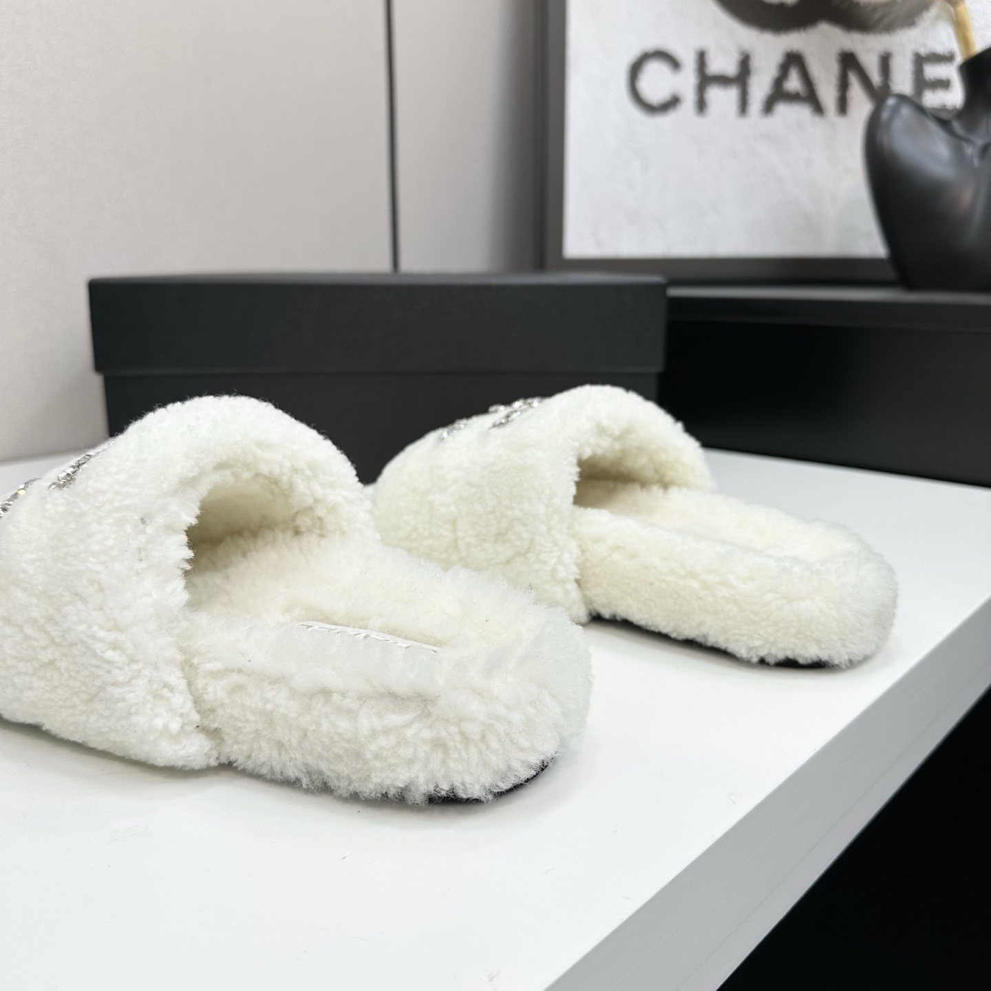 A pair of white fuzzy shearling slides with a crystal-embellished logo on a black box. - Hình 7