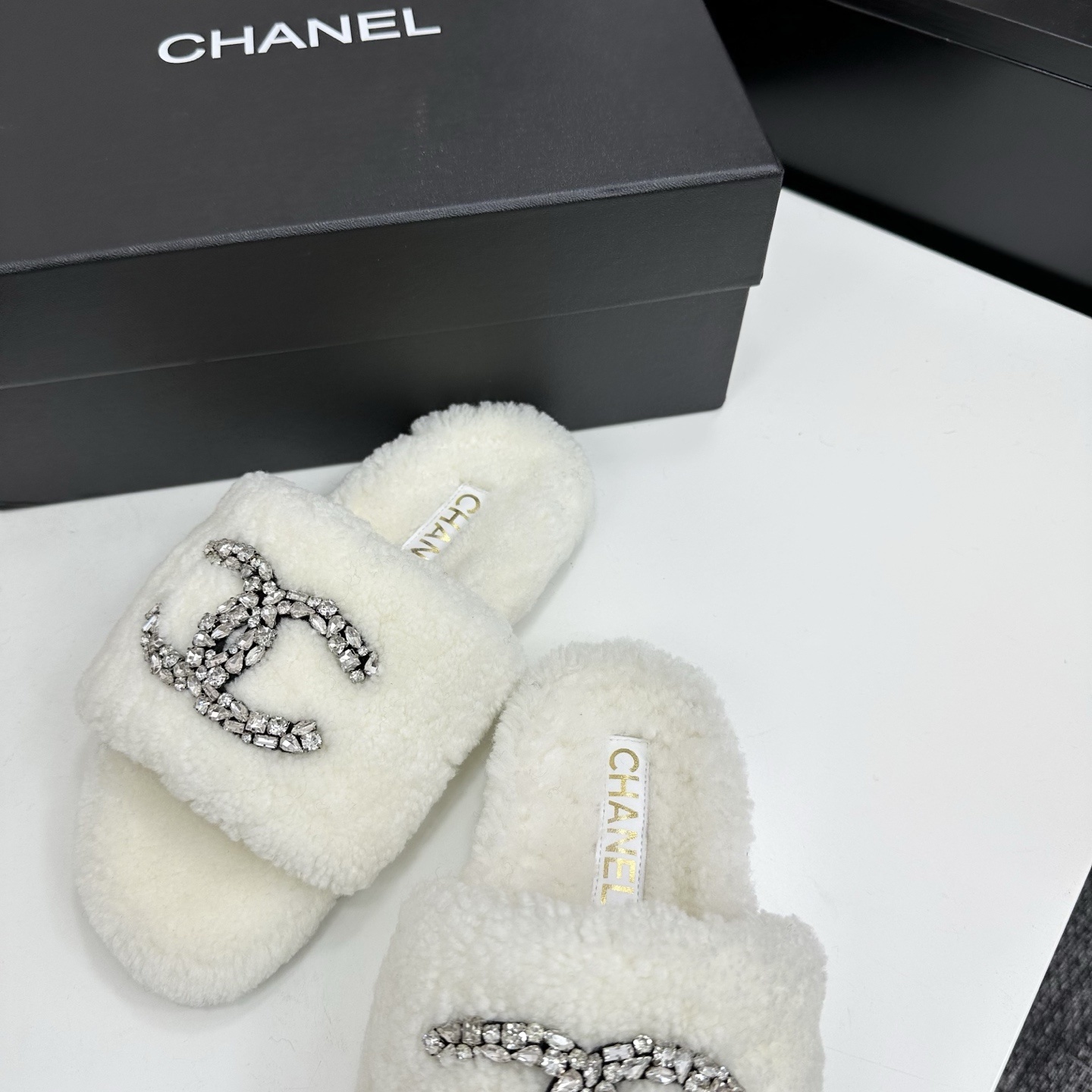 A pair of white fuzzy shearling slides with a crystal-embellished logo on a black box. - Hình 6