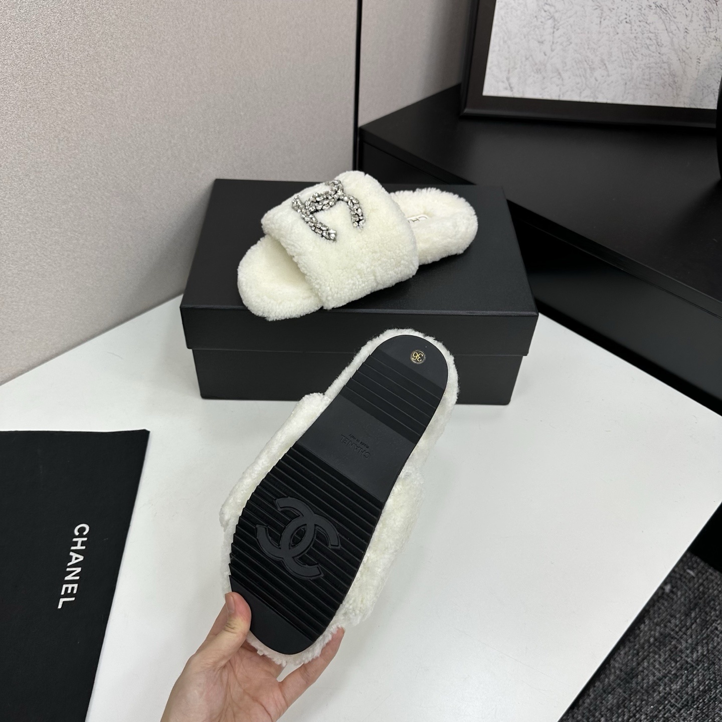 A pair of white fuzzy shearling slides with a crystal-embellished logo on a black box. - Hình 4