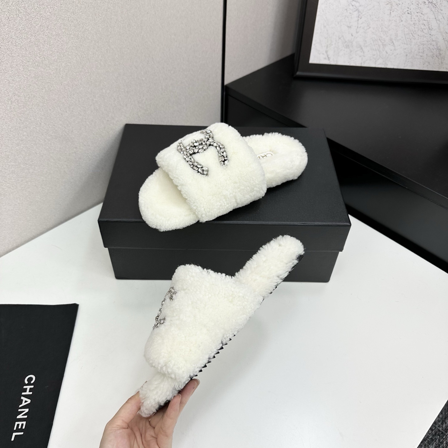 A pair of white fuzzy shearling slides with a crystal-embellished logo on a black box. - Hình 3