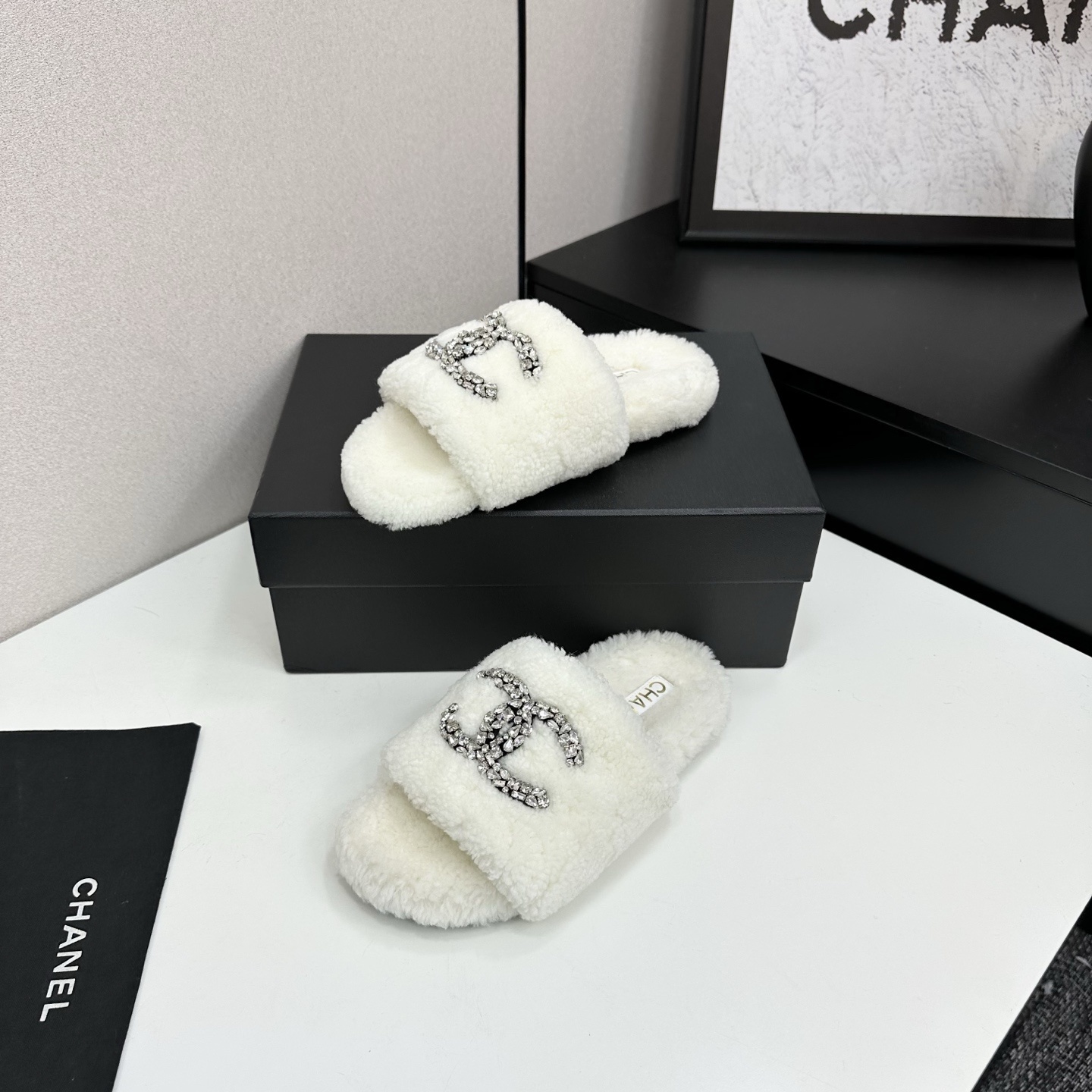A pair of white fuzzy shearling slides with a crystal-embellished logo on a black box. - Hình 2