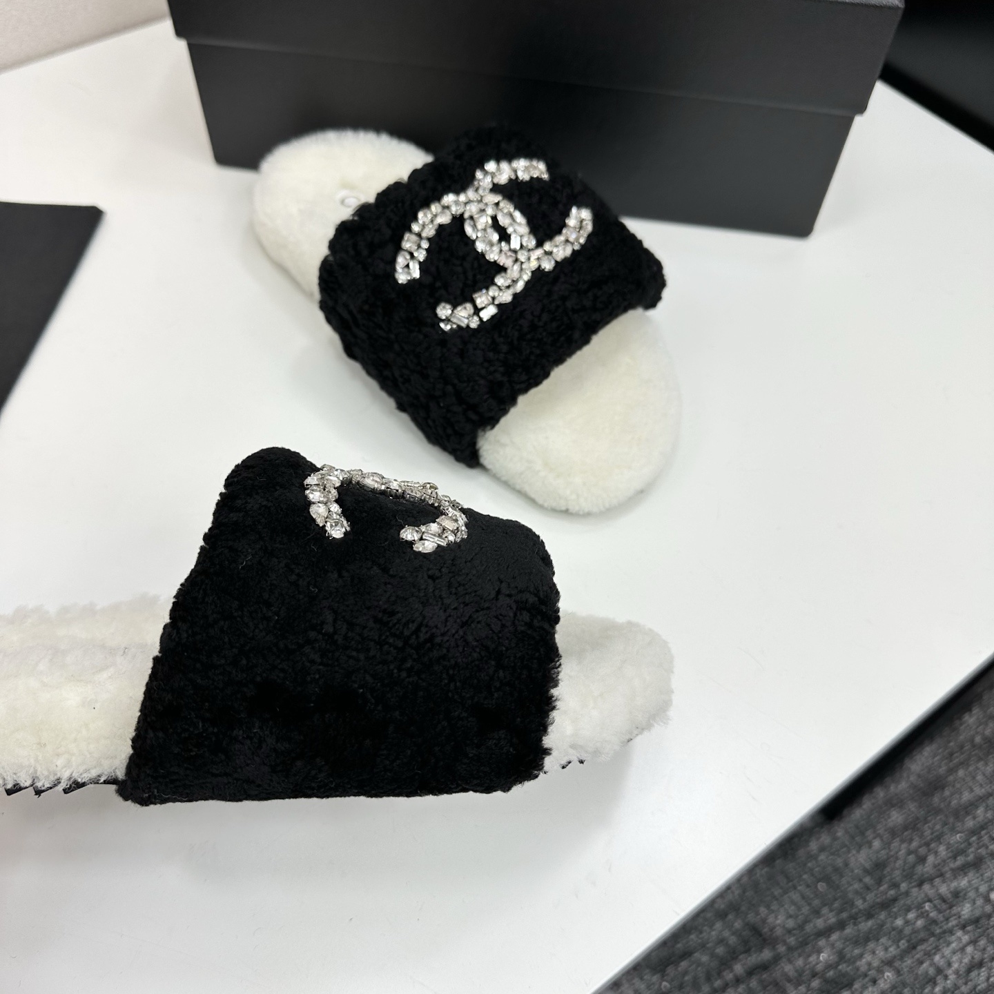 A pair of luxury black and white fuzzy slippers with a sparkling crystal-encrusted logo displayed on a black designer box. - Hình 9