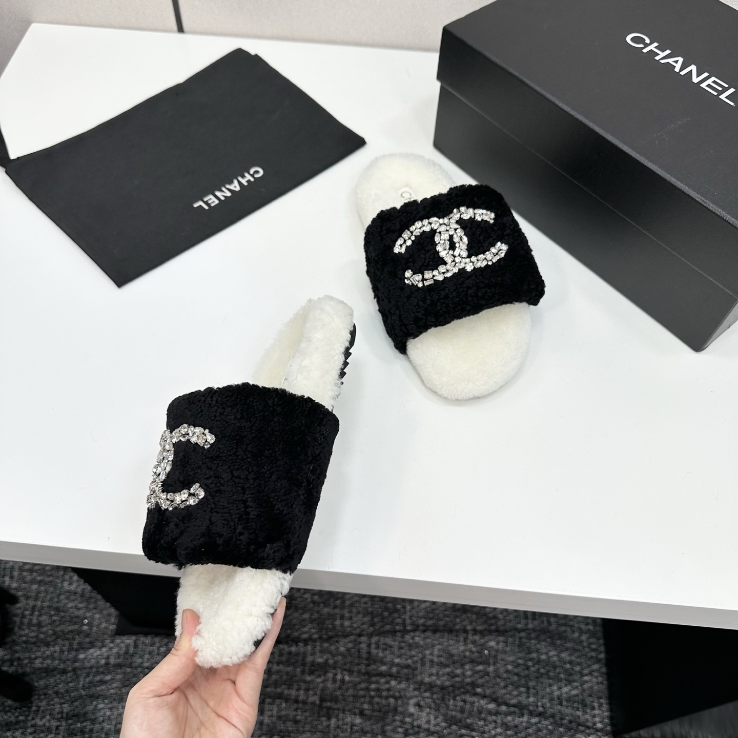 A pair of luxury black and white fuzzy slippers with a sparkling crystal-encrusted logo displayed on a black designer box. - Hình 8