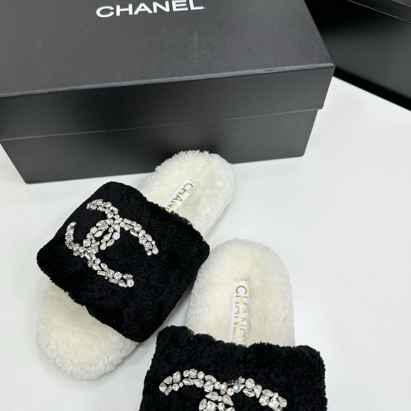 A pair of luxury black and white fuzzy slippers with a sparkling crystal-encrusted logo displayed on a black designer box. - Hình 6