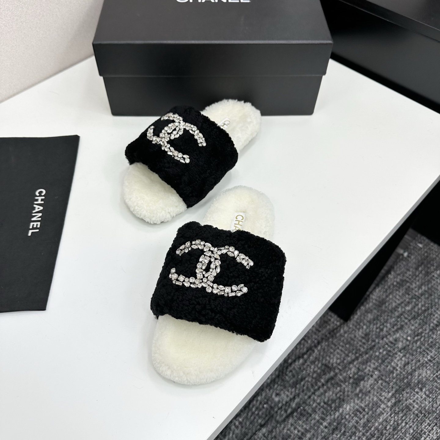 A pair of luxury black and white fuzzy slippers with a sparkling crystal-encrusted logo displayed on a black designer box. - Hình 5