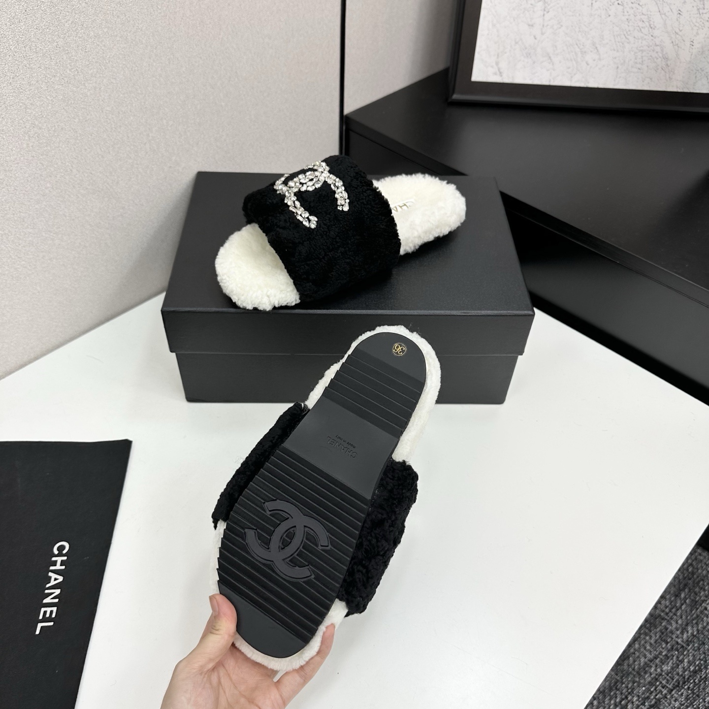 A pair of luxury black and white fuzzy slippers with a sparkling crystal-encrusted logo displayed on a black designer box. - Hình 4