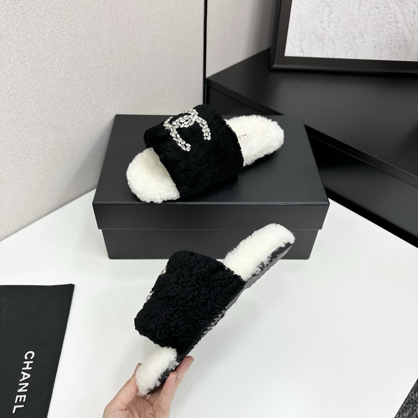 A pair of luxury black and white fuzzy slippers with a sparkling crystal-encrusted logo displayed on a black designer box. - Hình 3