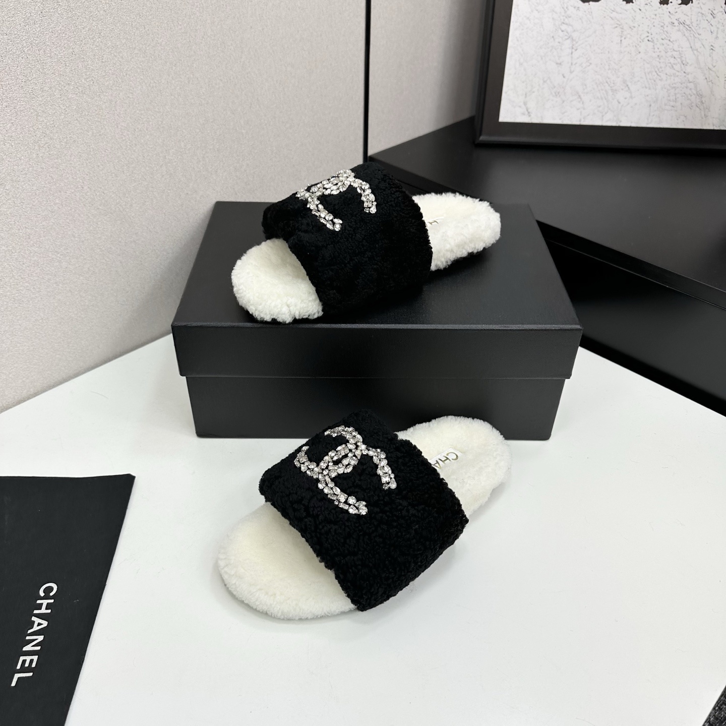 A pair of luxury black and white fuzzy slippers with a sparkling crystal-encrusted logo displayed on a black designer box. - Hình 2
