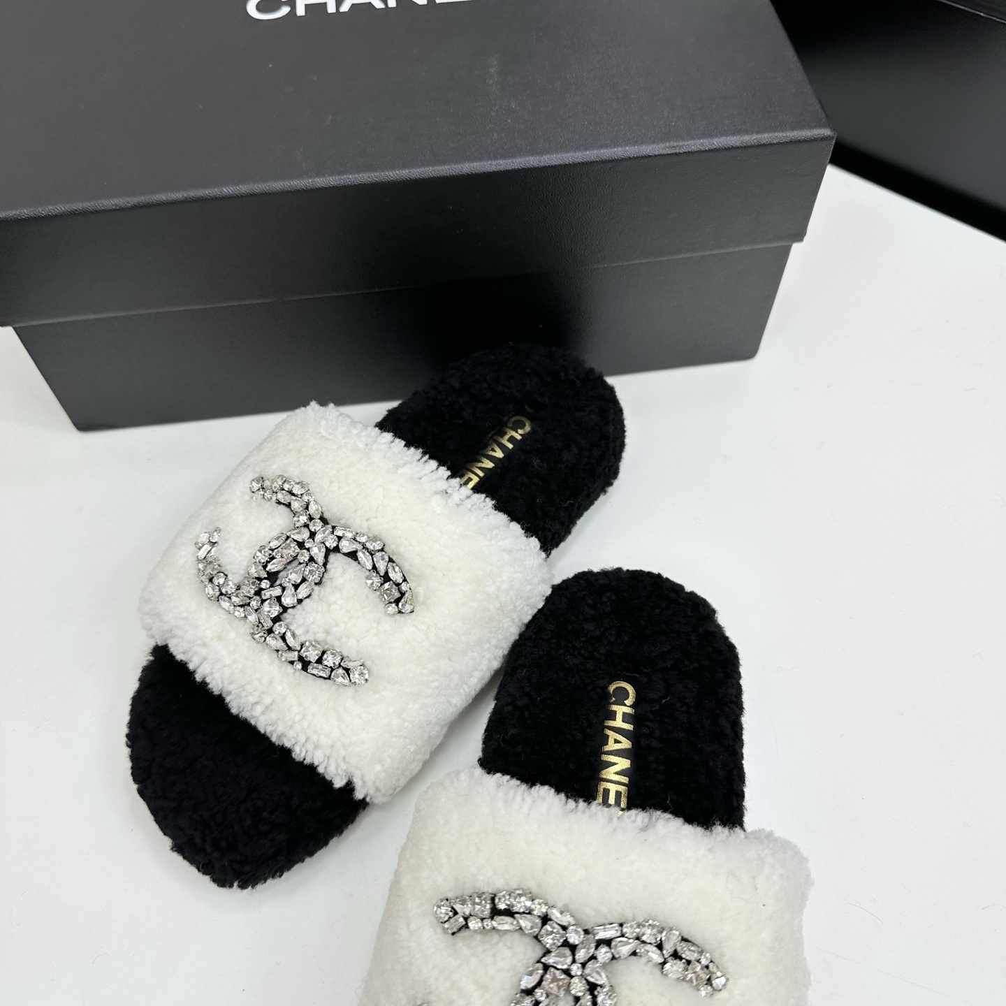 Pair of black and white fluffy shearling slides with a crystal-embellished interlocking logo sitting on a black box - Hình 6