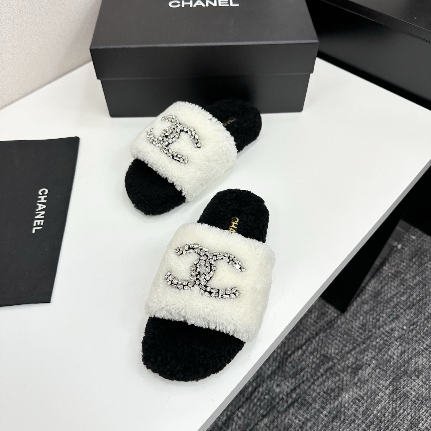 Pair of black and white fluffy shearling slides with a crystal-embellished interlocking logo sitting on a black box - Hình 5