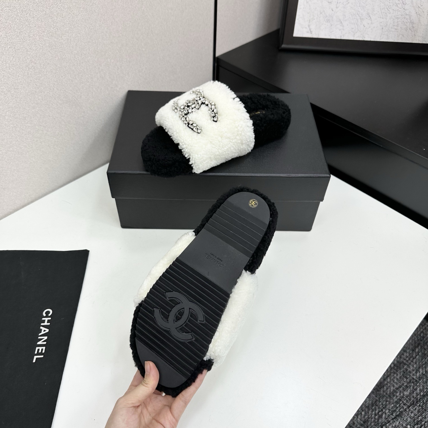 Pair of black and white fluffy shearling slides with a crystal-embellished interlocking logo sitting on a black box - Hình 4