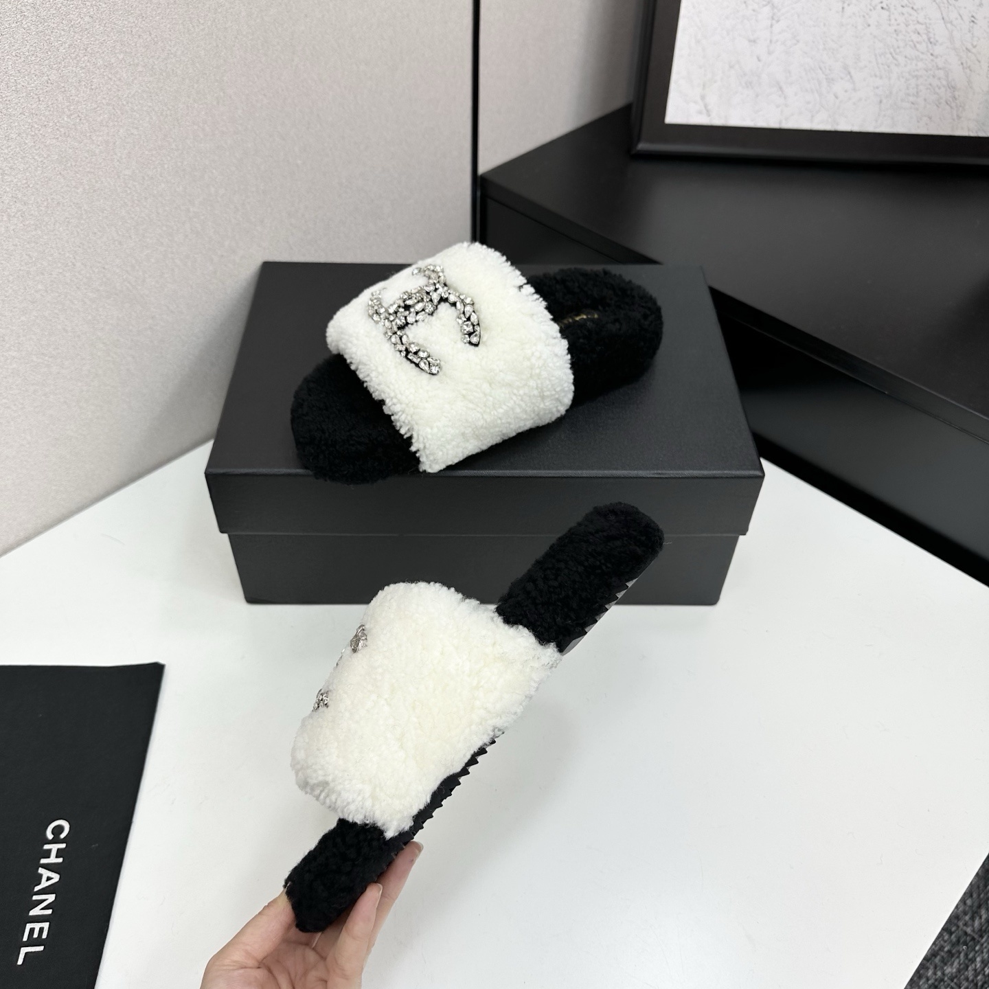 Pair of black and white fluffy shearling slides with a crystal-embellished interlocking logo sitting on a black box - Hình 3