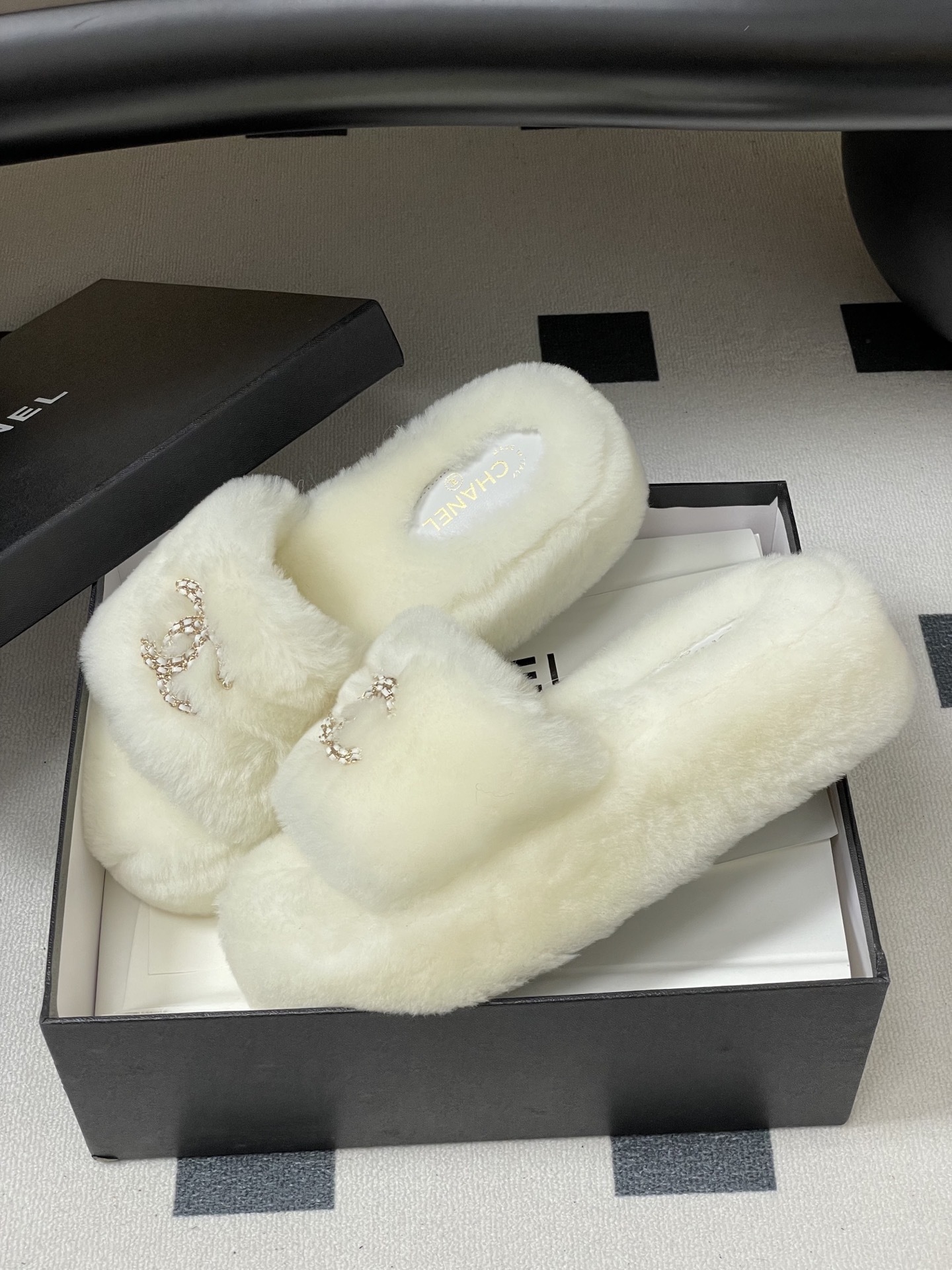 Pair of luxury Chanel white shearling fur slides with gold interlocking CC logo on a black surface. - Hình 4