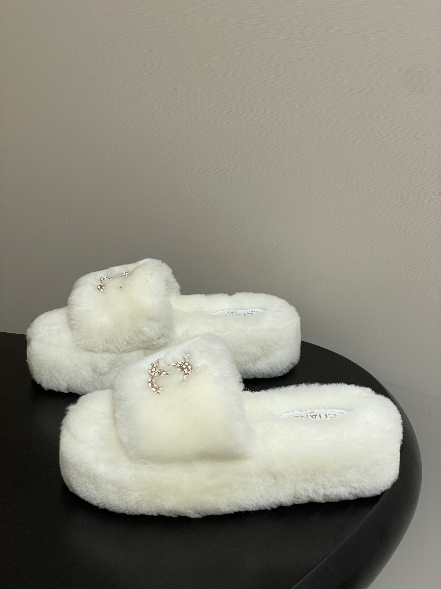 Pair of luxury Chanel white shearling fur slides with gold interlocking CC logo on a black surface. - Hình 3