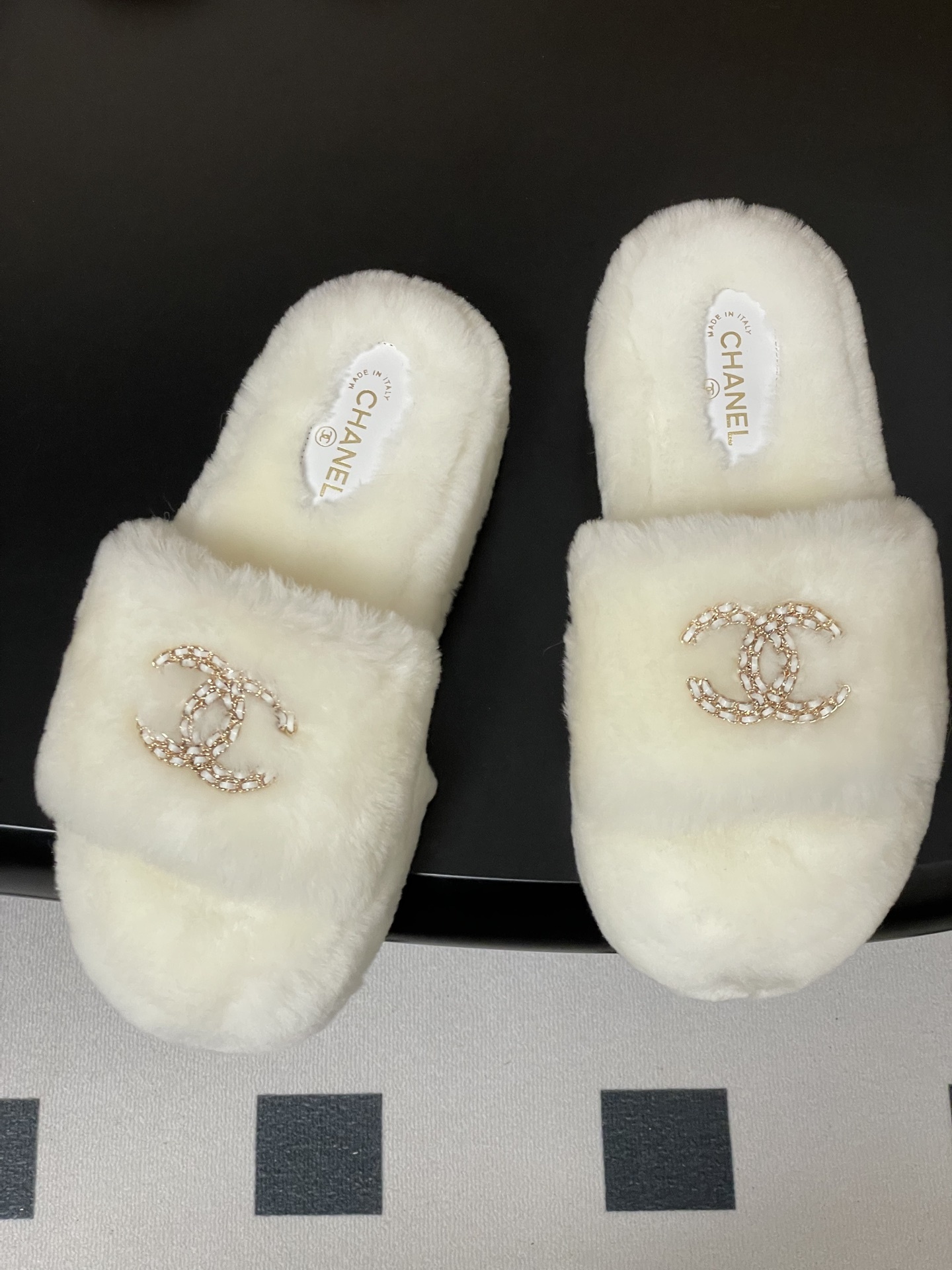 Pair of luxury Chanel white shearling fur slides with gold interlocking CC logo on a black surface. - Hình 2