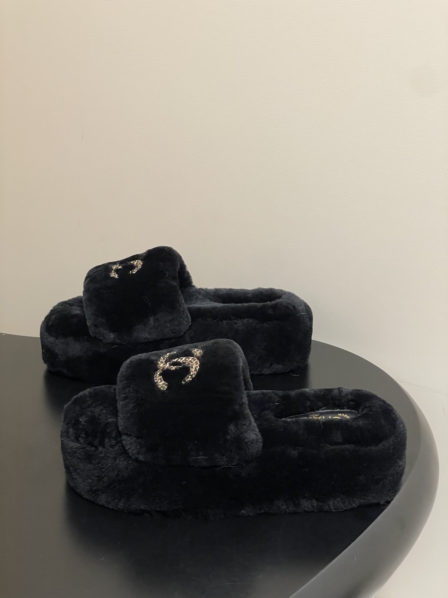 Pair of black Chanel shearling platform slides with gold chain interlocking CC logo on a black table. - Hình 3