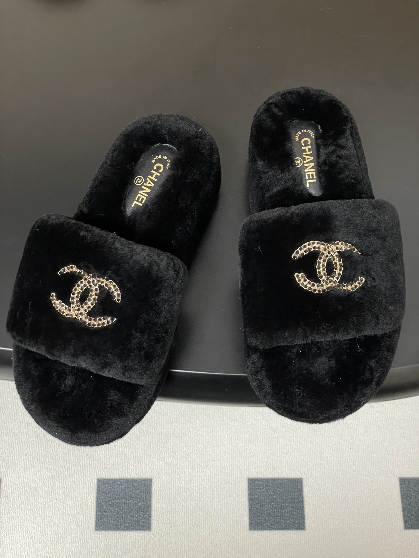 Pair of black Chanel shearling platform slides with gold chain interlocking CC logo on a black table. - Hình 2