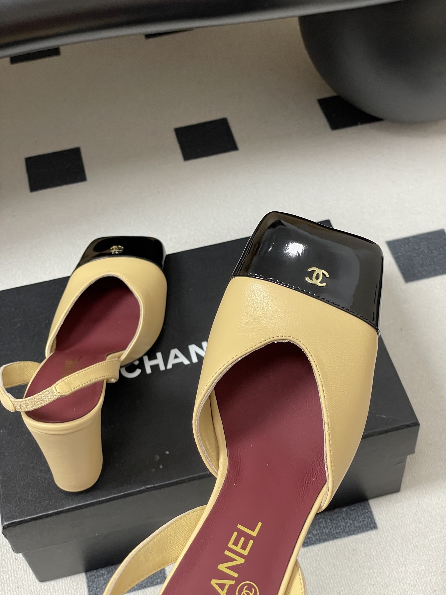 A pair of Chanel two-tone beige and black square toe slingback heels with block heels and gold CC logo on a black surface. - Hình 9