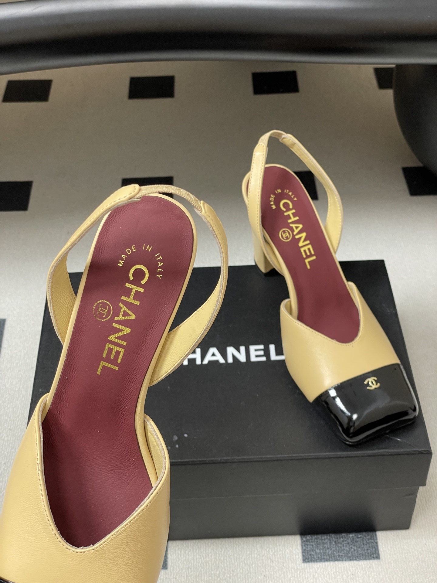 A pair of Chanel two-tone beige and black square toe slingback heels with block heels and gold CC logo on a black surface. - Hình 7