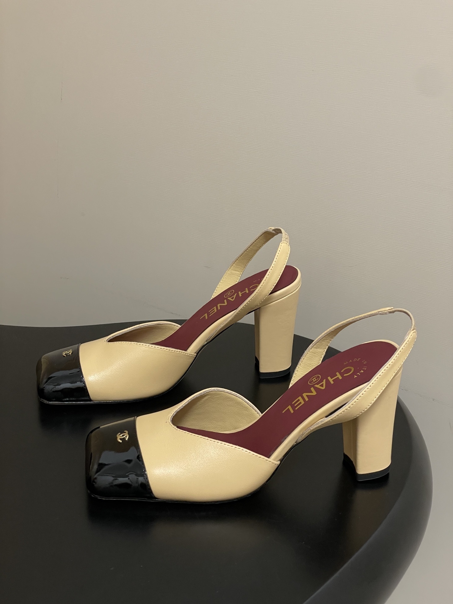 A pair of Chanel two-tone beige and black square toe slingback heels with block heels and gold CC logo on a black surface. - Hình 3