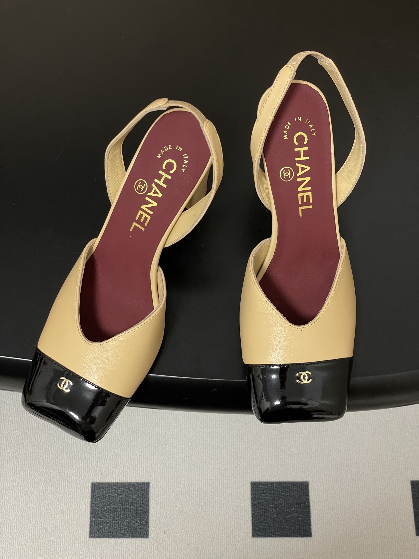 A pair of Chanel two-tone beige and black square toe slingback heels with block heels and gold CC logo on a black surface. - Hình 2