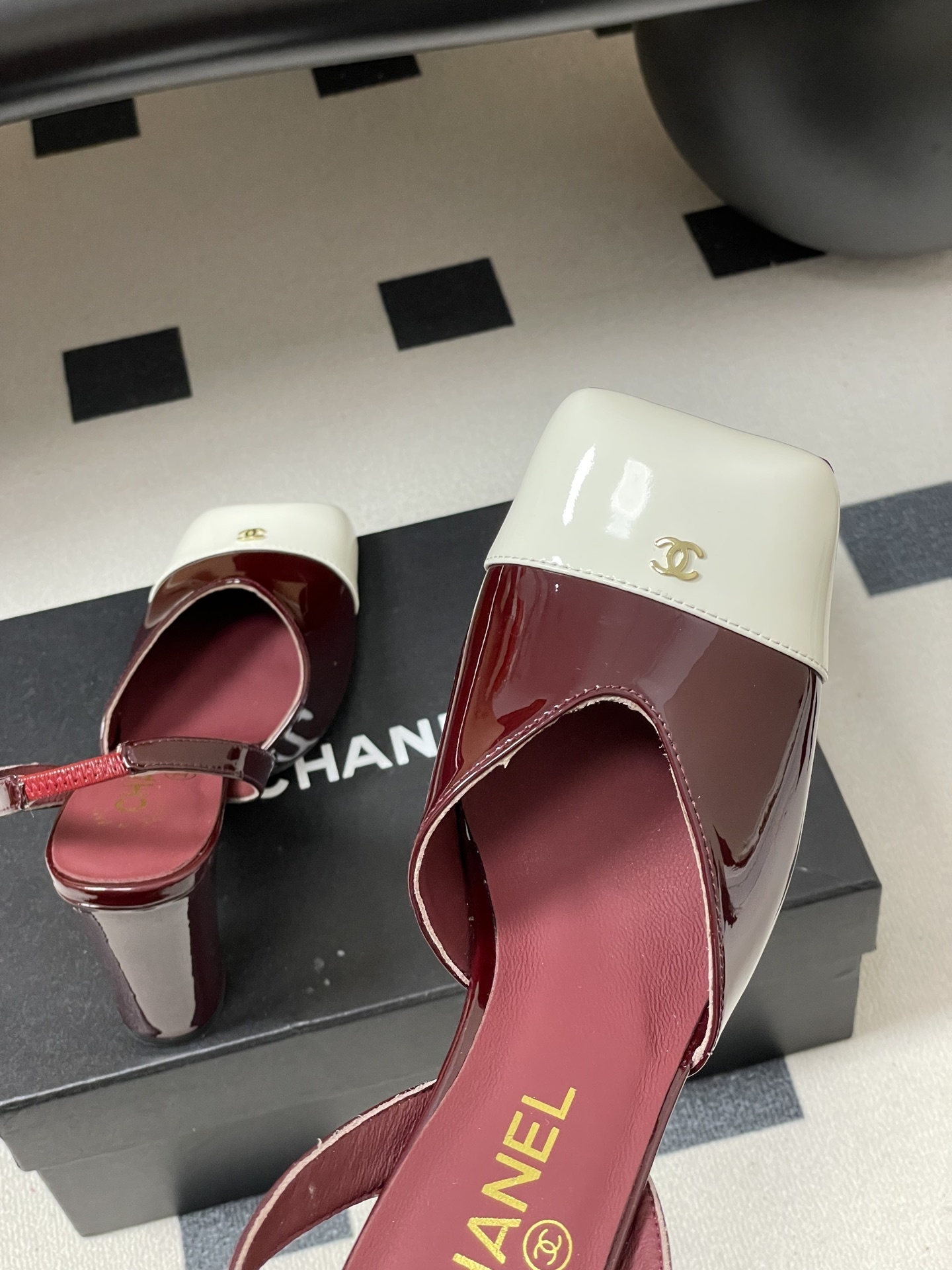 A pair of Chanel burgundy patent leather slingback pumps with a white square toe cap and gold CC logo on a black table. - Hình 9