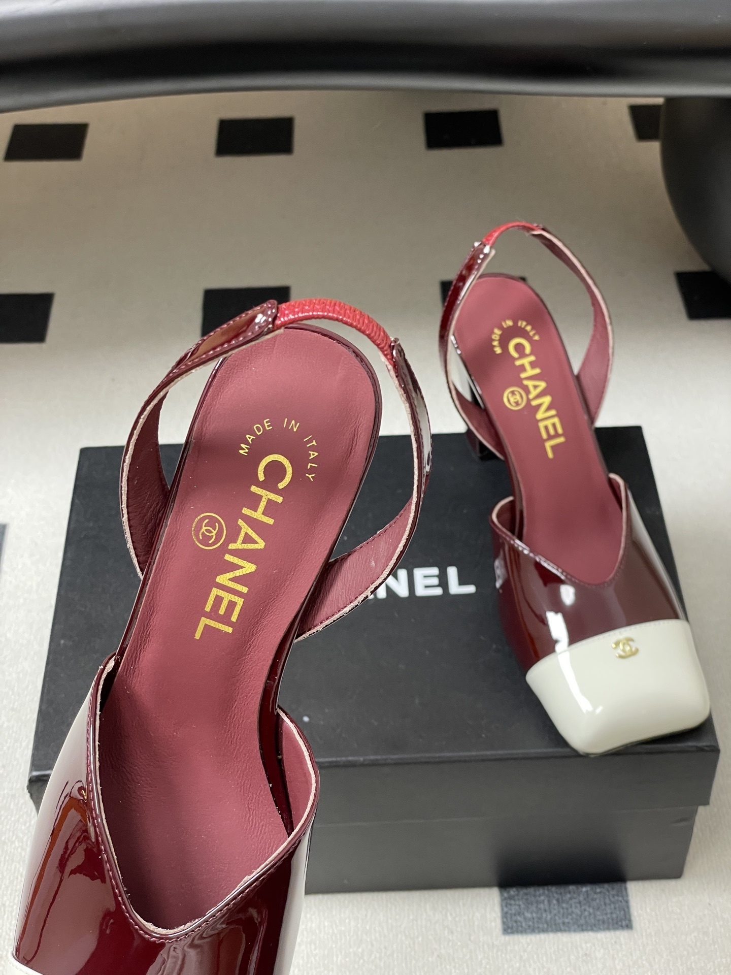 A pair of Chanel burgundy patent leather slingback pumps with a white square toe cap and gold CC logo on a black table. - Hình 7