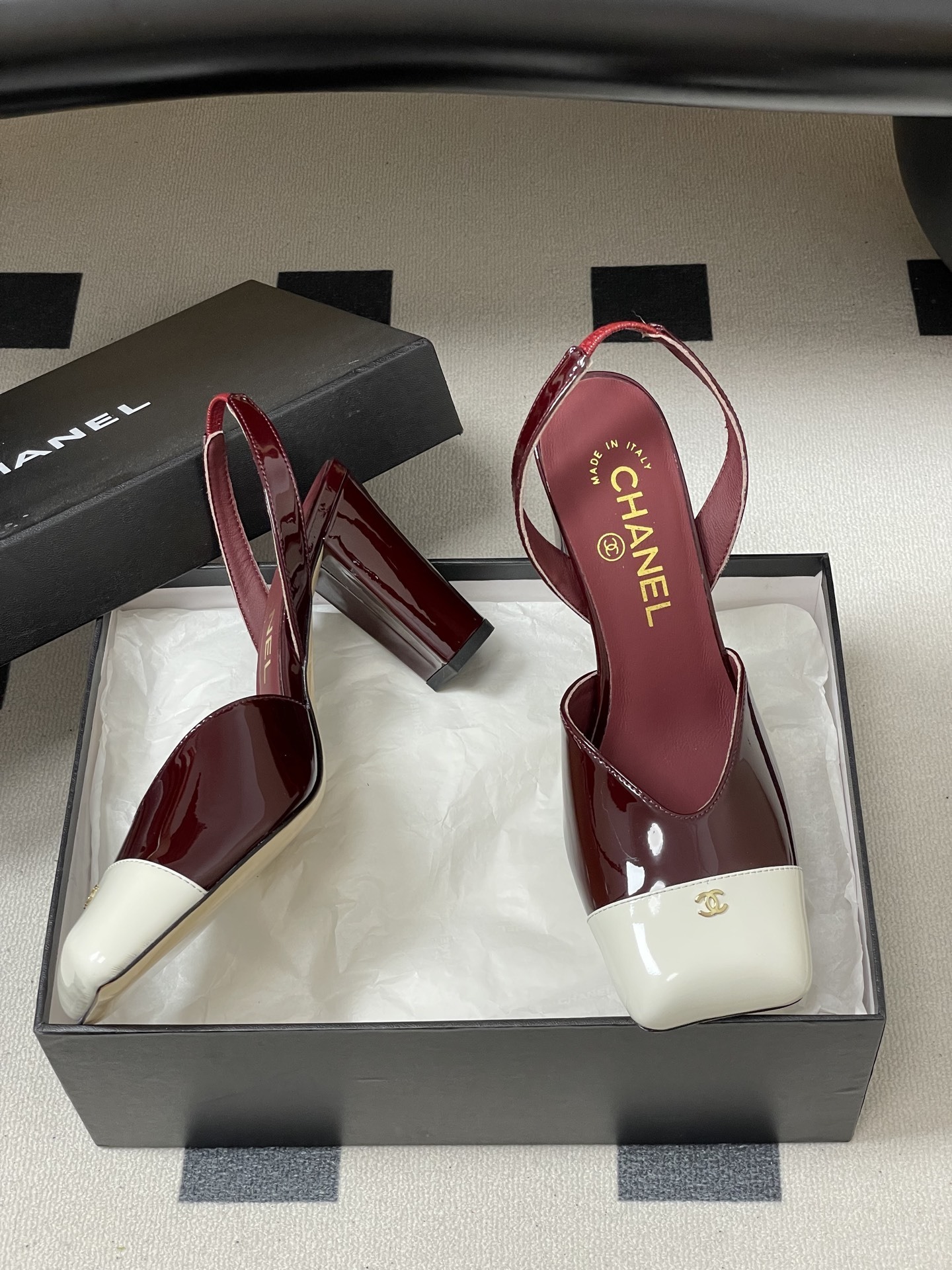 A pair of Chanel burgundy patent leather slingback pumps with a white square toe cap and gold CC logo on a black table. - Hình 6