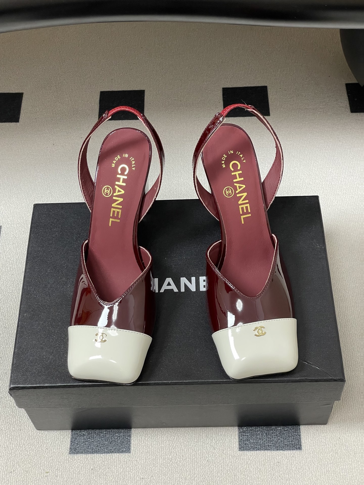 A pair of Chanel burgundy patent leather slingback pumps with a white square toe cap and gold CC logo on a black table. - Hình 5