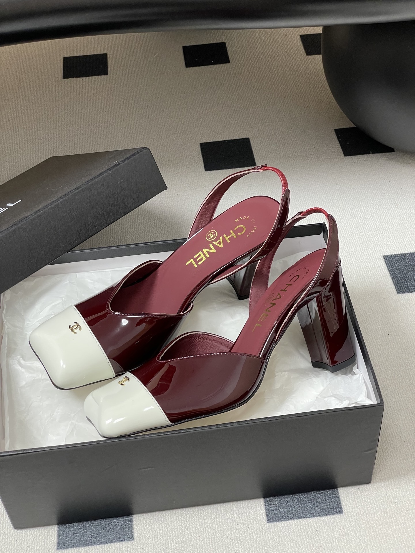 A pair of Chanel burgundy patent leather slingback pumps with a white square toe cap and gold CC logo on a black table. - Hình 4