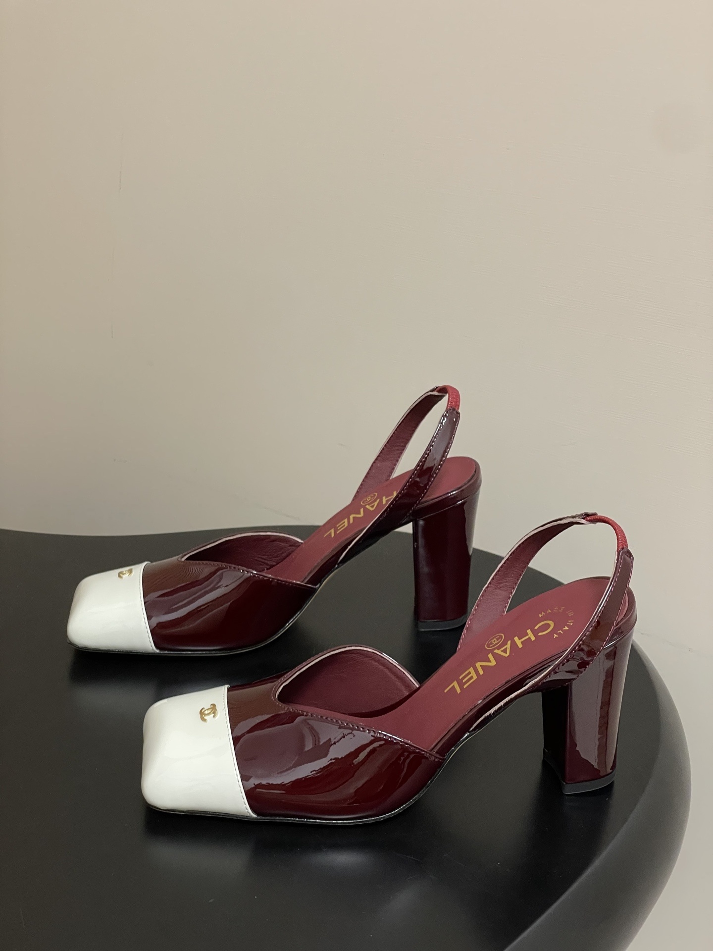 A pair of Chanel burgundy patent leather slingback pumps with a white square toe cap and gold CC logo on a black table. - Hình 3