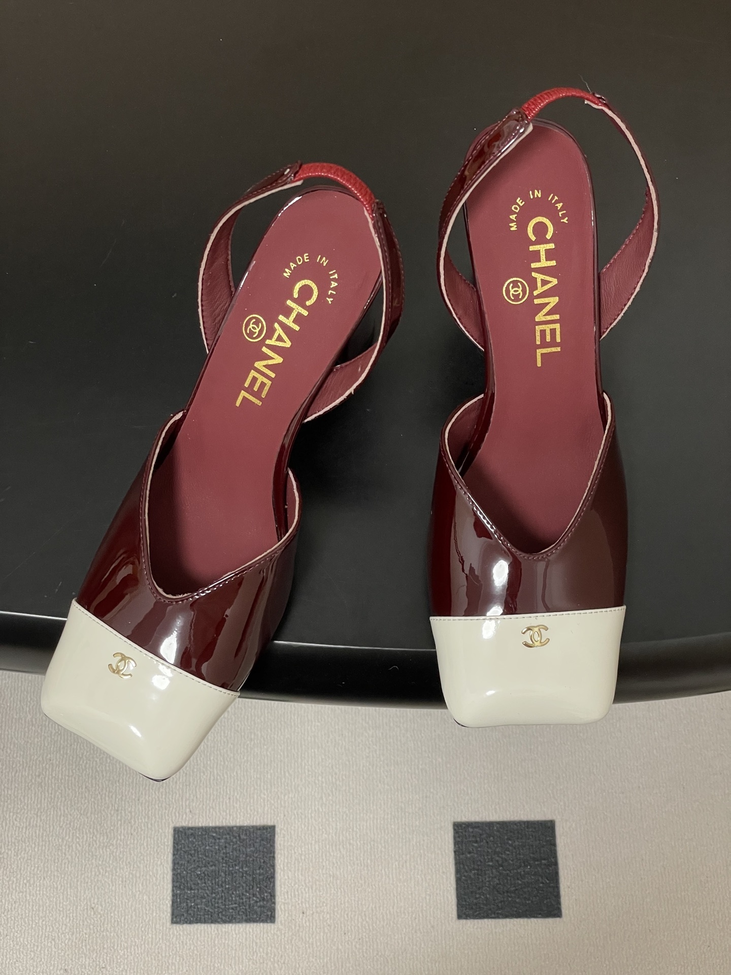 A pair of Chanel burgundy patent leather slingback pumps with a white square toe cap and gold CC logo on a black table. - Hình 2