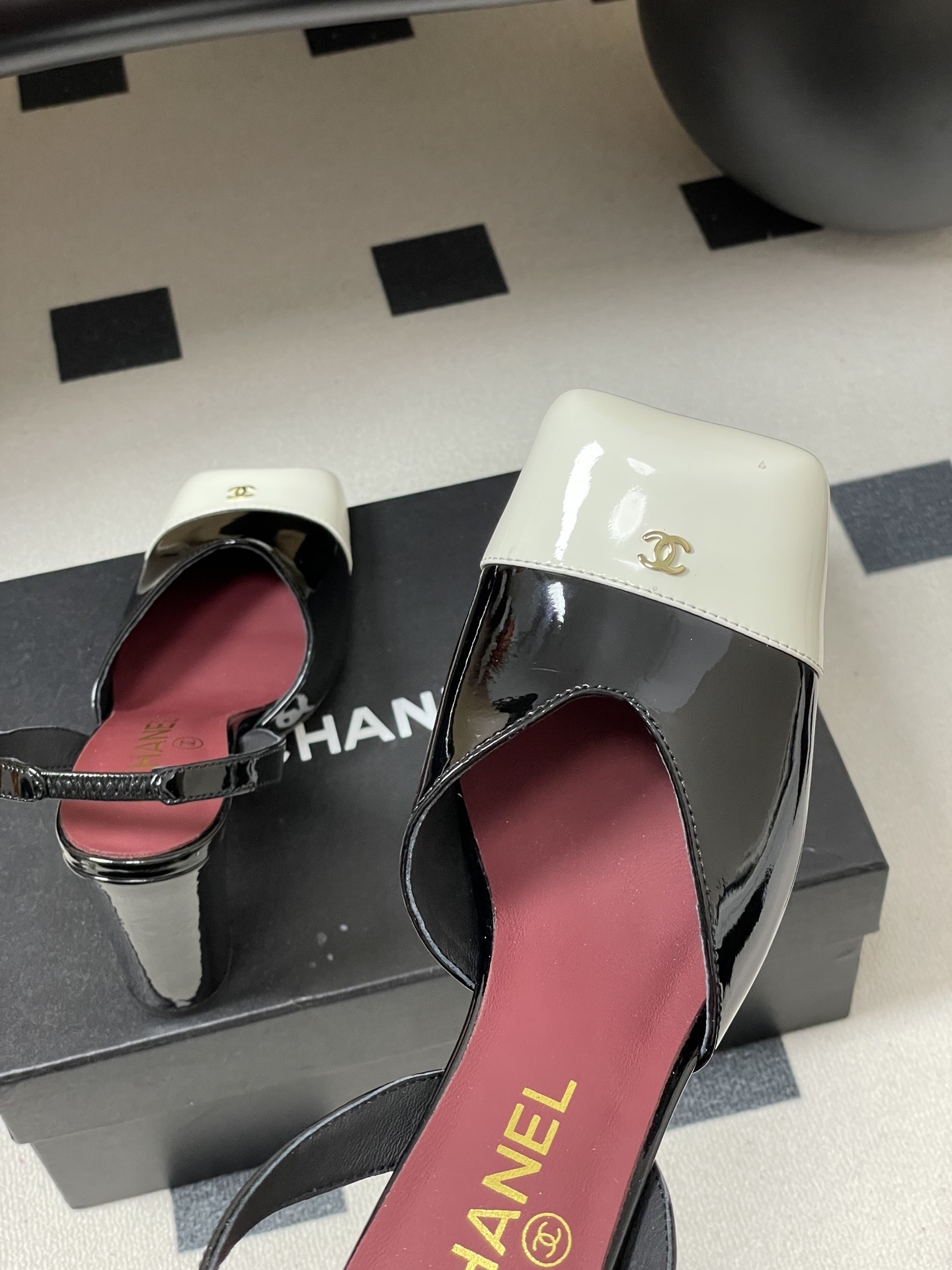 Pair of Chanel black patent leather slingback heels with white square toe caps and gold CC logo on a black table. - Hình 9