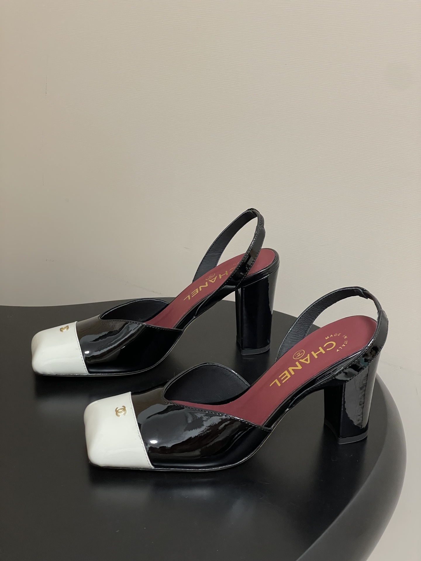 Pair of Chanel black patent leather slingback heels with white square toe caps and gold CC logo on a black table. - Hình 3