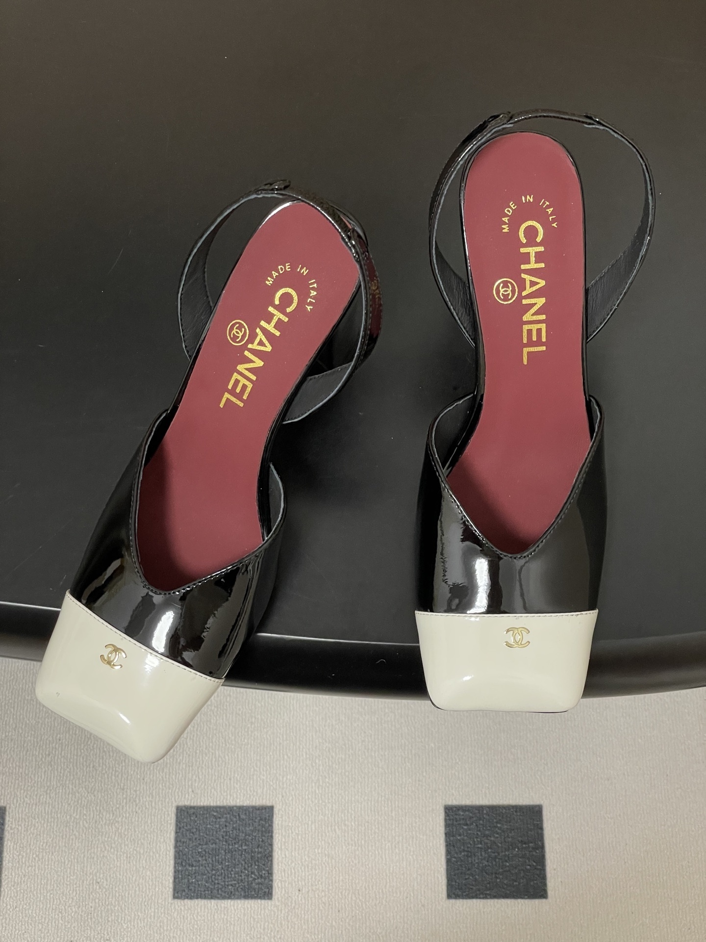 Pair of Chanel black patent leather slingback heels with white square toe caps and gold CC logo on a black table. - Hình 2