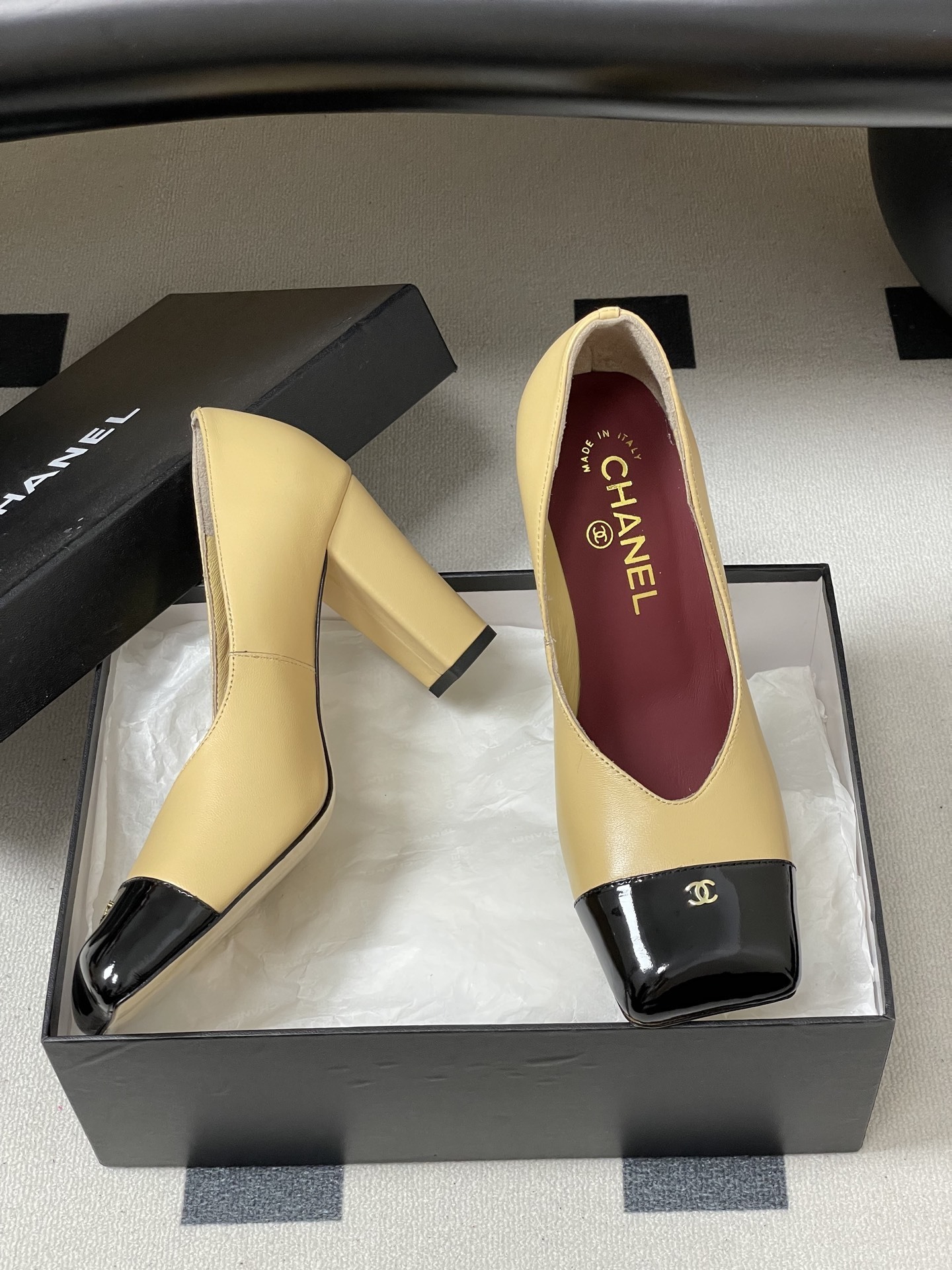 A pair of Chanel beige leather pumps featuring a black patent leather square toe cap with gold CC logo and a sturdy block heel. - Hình 6