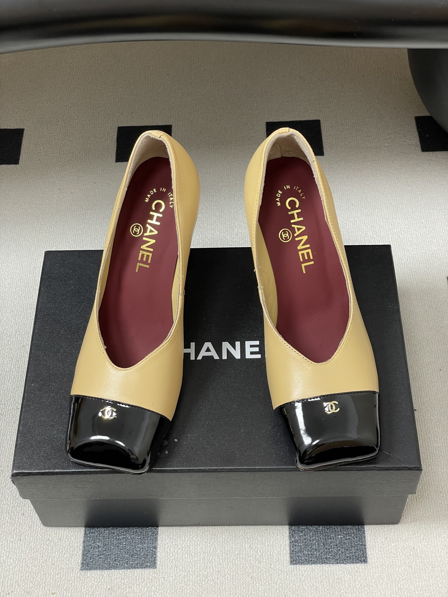 A pair of Chanel beige leather pumps featuring a black patent leather square toe cap with gold CC logo and a sturdy block heel. - Hình 5