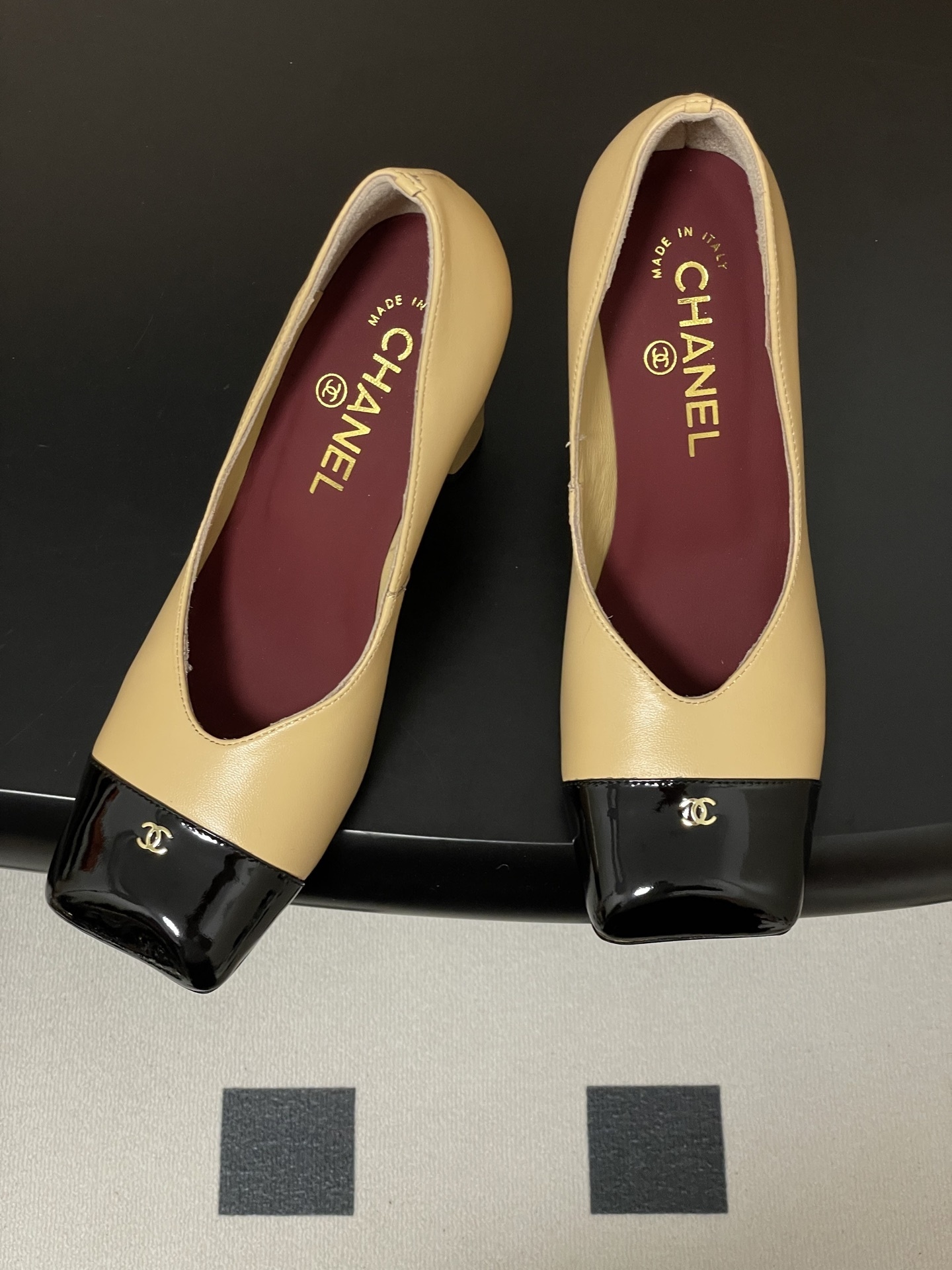 A pair of Chanel beige leather pumps featuring a black patent leather square toe cap with gold CC logo and a sturdy block heel. - Hình 2