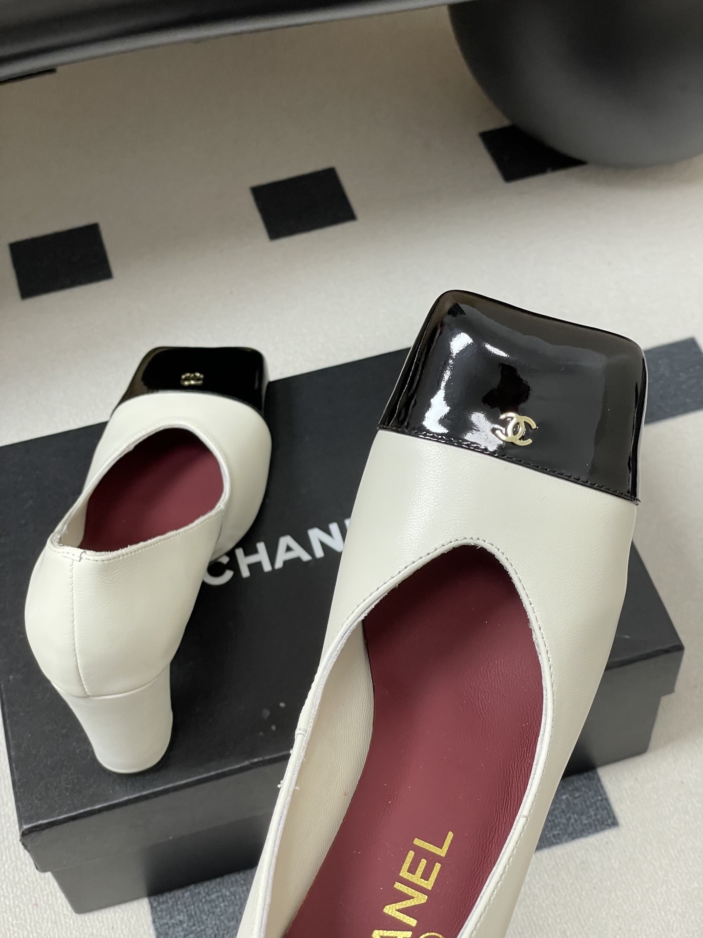 A pair of Chanel two-tone block heel pumps in white leather with black patent square toes and gold CC logos on a black table. - Hình 9