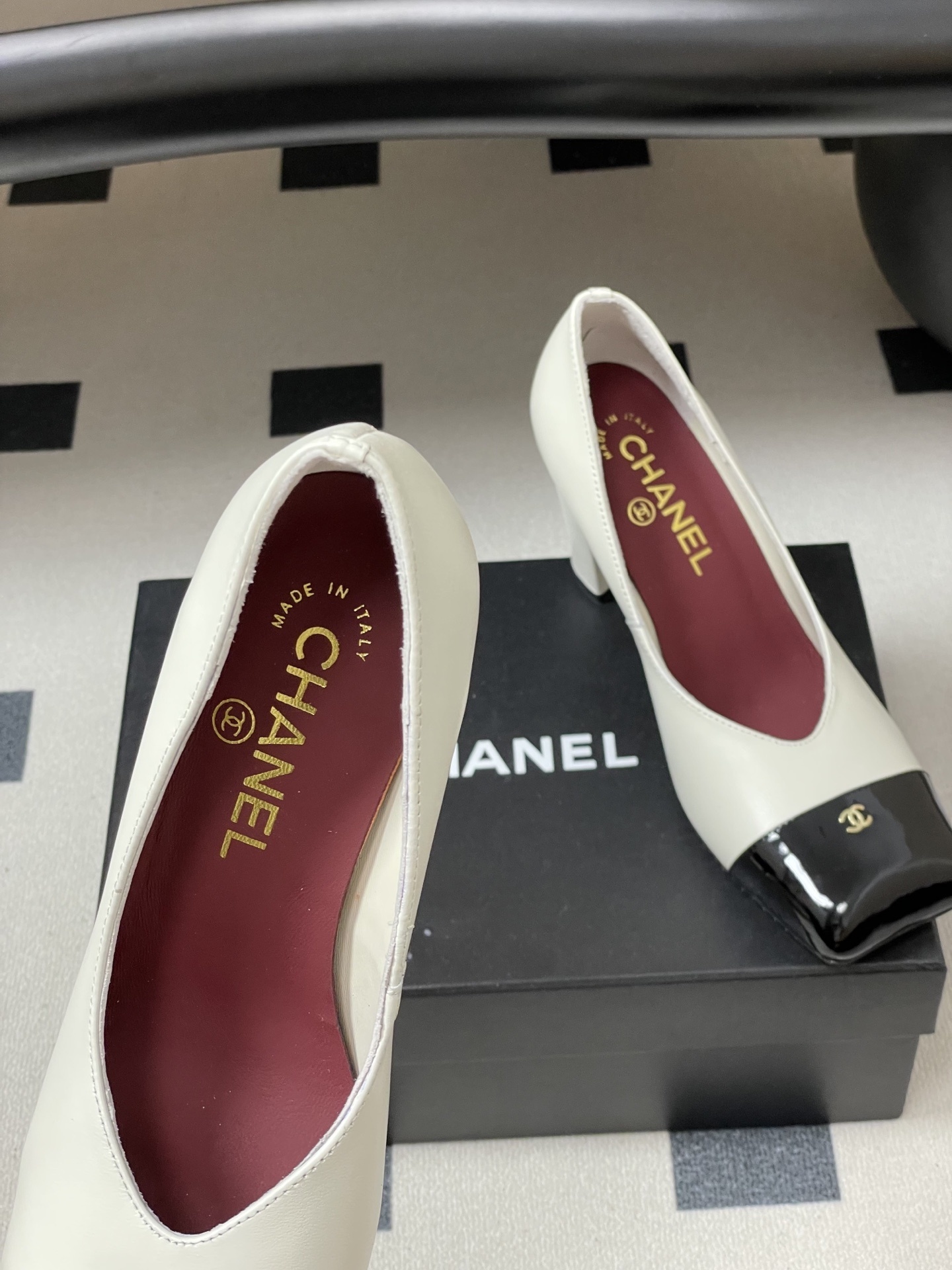 A pair of Chanel two-tone block heel pumps in white leather with black patent square toes and gold CC logos on a black table. - Hình 7