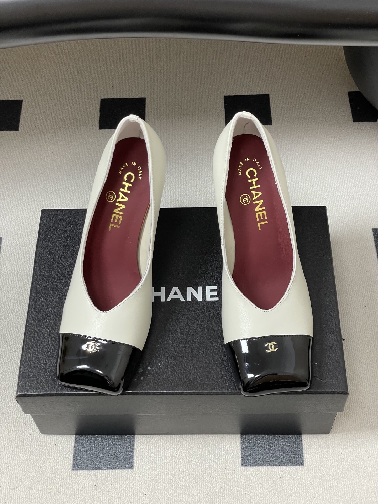 A pair of Chanel two-tone block heel pumps in white leather with black patent square toes and gold CC logos on a black table. - Hình 5